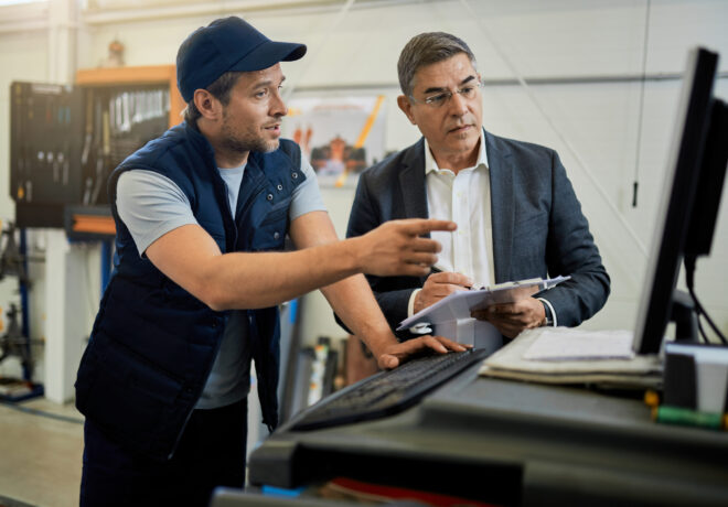Mid adult manager and car mechanic talking while using computer in auto repair shop.